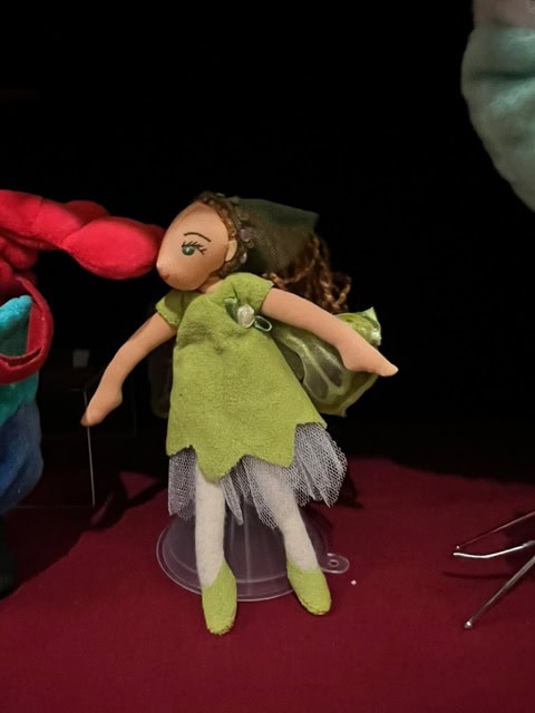 a cloth doll dressed as a fairy with green wings, a green dress, and white leggings stands on a red surface against a dark background.