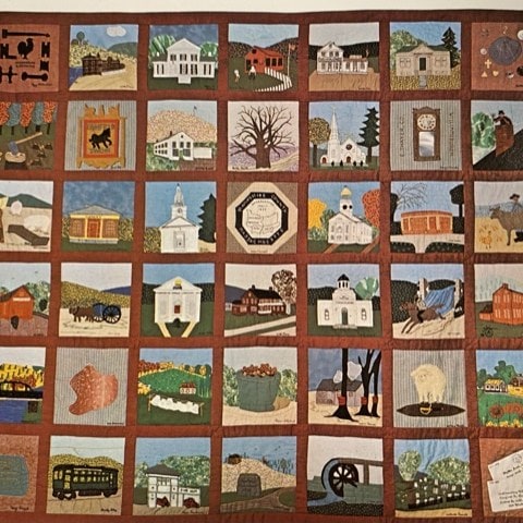 a handmade quilt featuring various squares, each depicting a different building, landscape, or local scene, bordered by brown sashing.