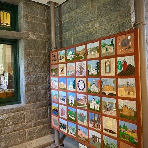 a colorful quilt featuring various buildings and scenes hangs on a wall next to a window in a stone walled room.