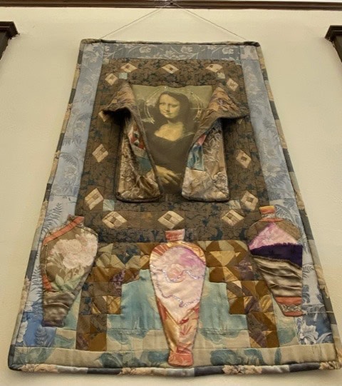 a quilted wall hanging features the mona lisa at the top center with three fabric vases below, surrounded by patterned patchwork in blue, brown, and tan tones.
