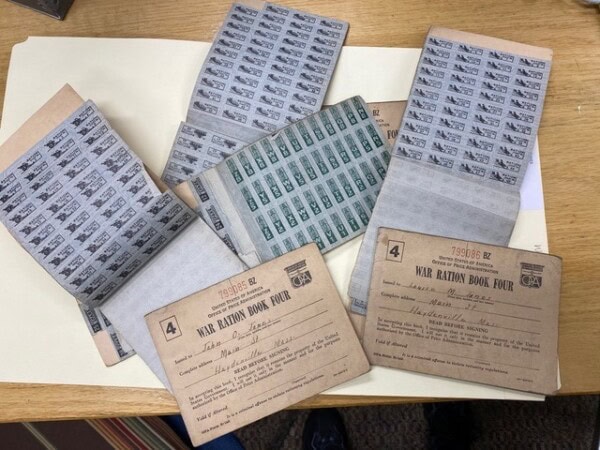 several wwii era war ration books and sheets of ration stamps are displayed on a wooden table, with handwritten information visible on the book covers.