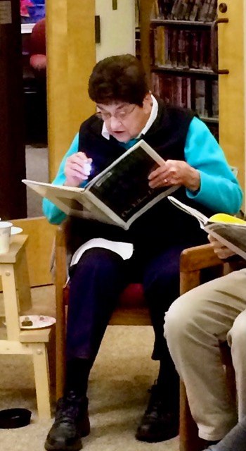 an older woman sits in a chair reading a book, using a small flashlight. bookshelves and another seated person are visible in the background.