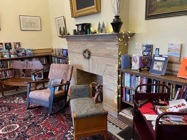 a cozy library corner with two chairs, a patterned rug, a fireplace decorated with a wreath, shelves of books, and a cart holding papers and books.