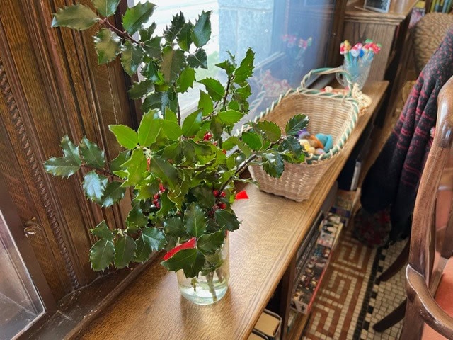 a glass vase with holly branches sits on a wooden windowsill next to a wicker basket filled with small balls of yarn and other craft items.