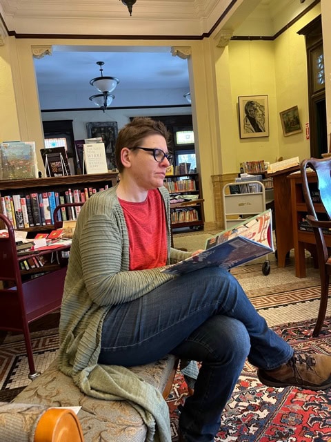 a person with glasses sits cross legged on a bench in a library, reading a book. shelves of books and framed photos are visible in the background.