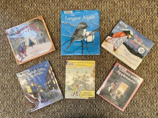 six children's holiday books are arranged on a carpeted floor, featuring winter and festive themes such as christmas, hanukkah, and applesauce.