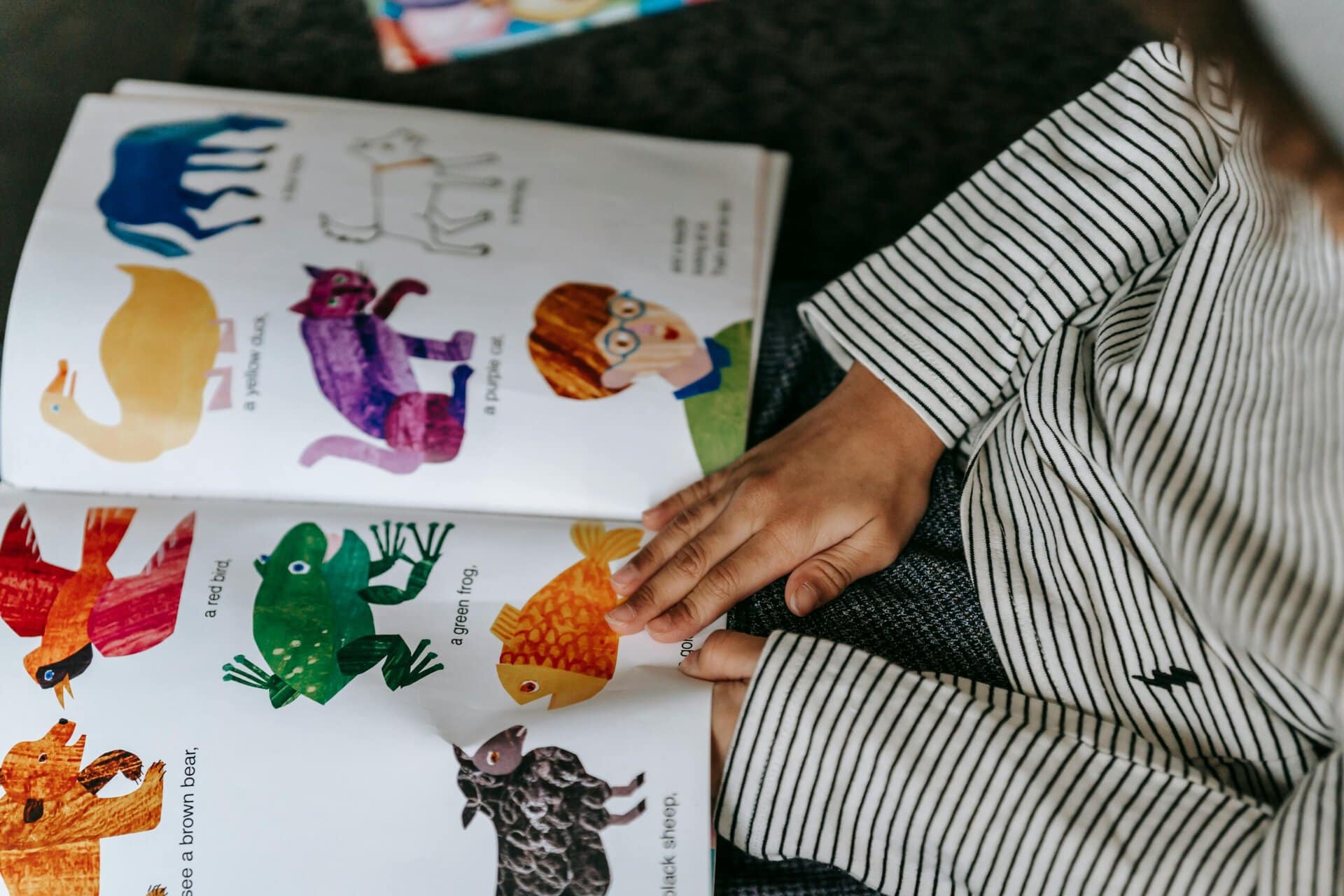 a child wearing a striped shirt reads a colorful illustrated animal book, showing pictures of a frog, fish, bear, and other animals.
