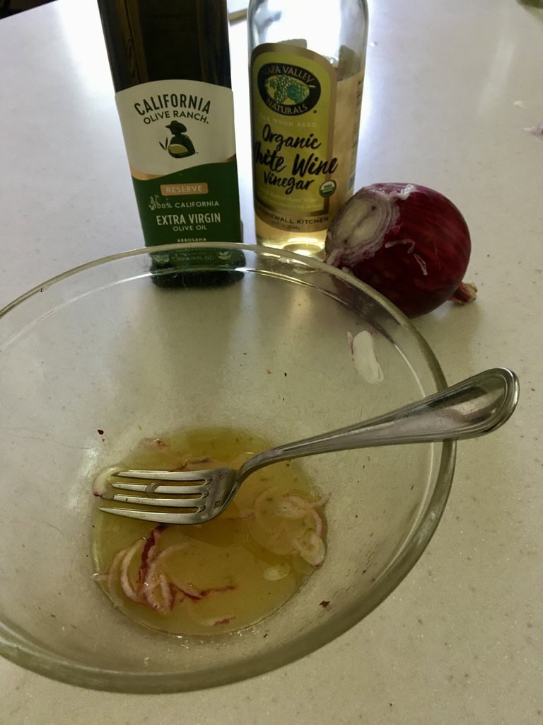 a glass bowl with a fork, sliced red onion, and dressing sits in front of bottles of olive oil and white wine vinegar on a countertop.