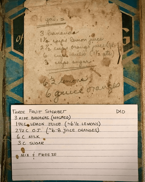 a handwritten recipe card for three fruit sherbet with ingredients and instructions is pinned over an old, stained sherbet recipe taped to a blue patterned book cover.