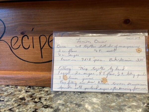 a handwritten recipe card for lemon bars sits in front of a wooden recipe box on a kitchen counter. the card lists ingredients and baking instructions.