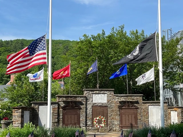 a memorial with a stone wall, wreath, and multiple flags, including the u.s. flag and military flags, displayed in front of green trees and a blue sky.