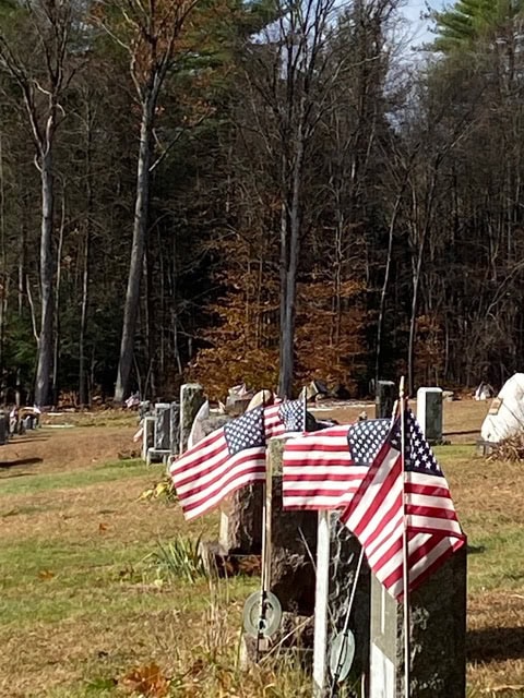 several gravestones marked with american flags stand in a cemetery surrounded by trees with autumn foliage.