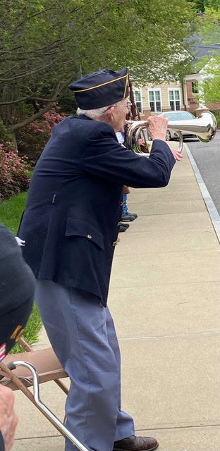 an elderly man in a military uniform plays a bugle while standing on a sidewalk in a residential neighborhood.