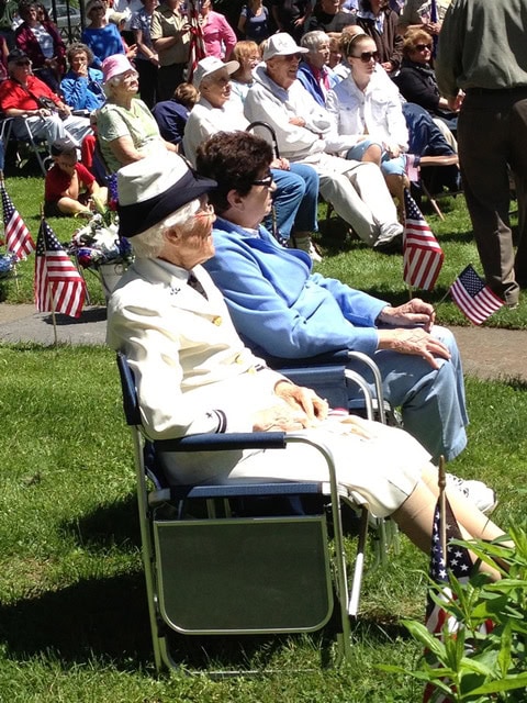 two elderly women sit in chairs outdoors at an event, surrounded by american flags and other seated attendees on a sunny day.