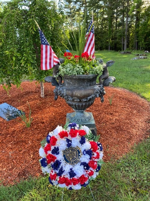 a decorative urn with flowers and two american flags sits on mulch, with a red, white, and blue wreath bearing a u.s. navy emblem placed in front. trees and grass are in the background.