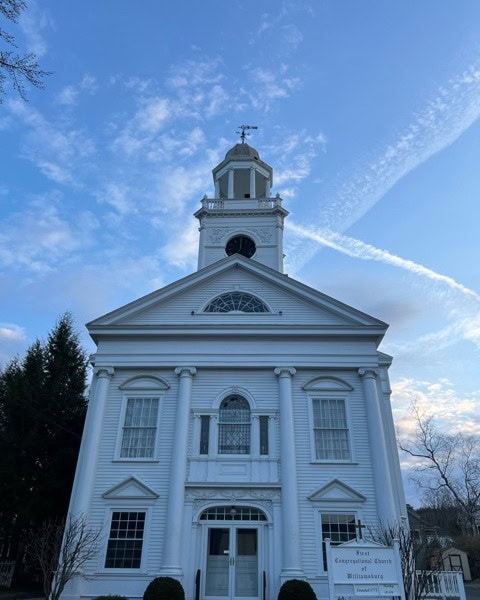 white church with a central clock tower and columns at the entrance, set against a partly cloudy blue sky with contrails.