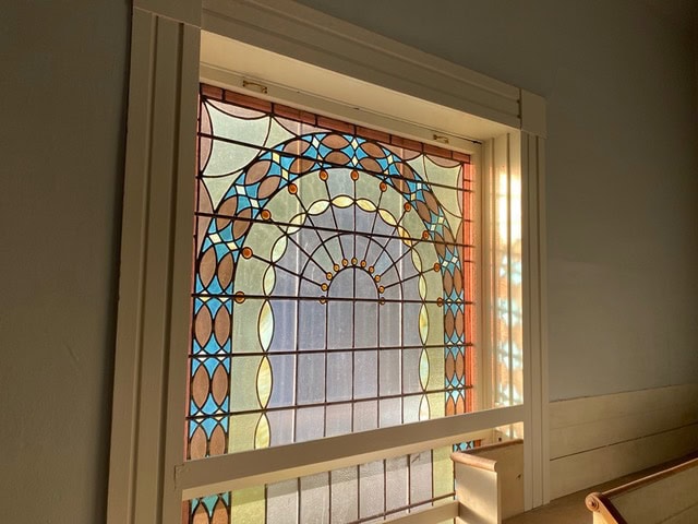 a stained glass window with geometric patterns in blue, gold, and brown is illuminated by sunlight from outside, framed by white trim.