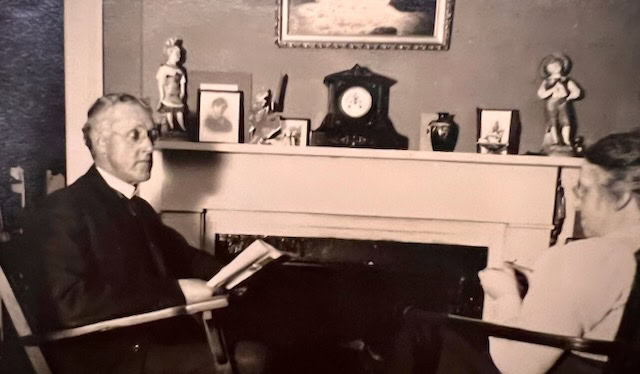 two people sit facing each other in a living room with a mantel adorned with a clock, framed photos, and figurines. one person holds a book.