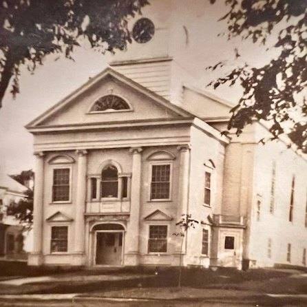 a sepia toned photo of a two story, historic church with large columns, arched windows, and a clock tower, partially framed by tree branches.