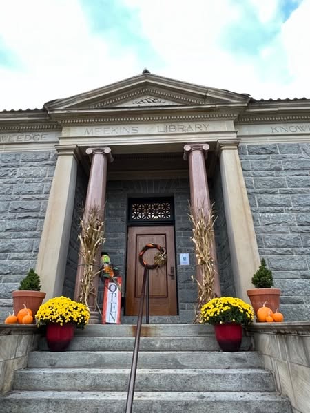 stone library entrance with two columns, fall decorations including yellow flowers, pumpkins, and cornstalks. a sign by the door reads "open." building labeled "meekins library.