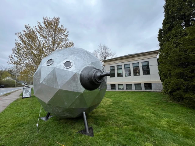 a large, silver, geometric sphere sculpture with circular windows and a protruding nozzle stands on metal legs outside a building on a grassy lawn.