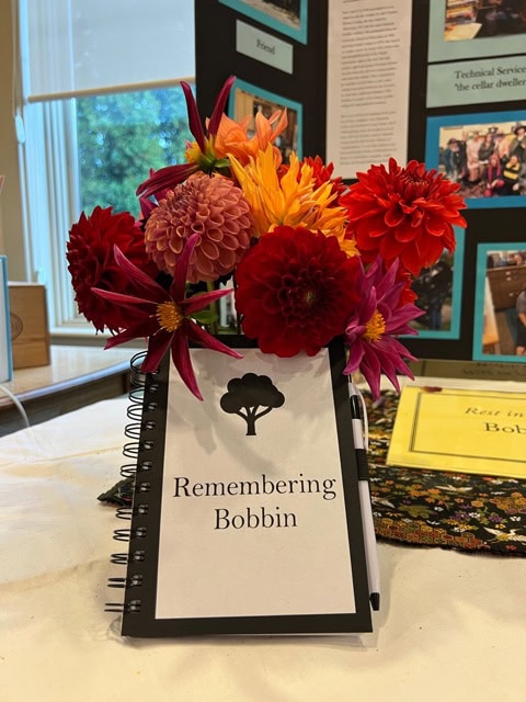 a spiral bound booklet titled "remembering bobbin" with a flower arrangement on top, set on a table with a photo display board in the background.