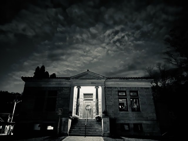 a stone building with columns is illuminated at night, with dramatic clouds in the sky above and a wheelchair accessible sign visible near the entrance.