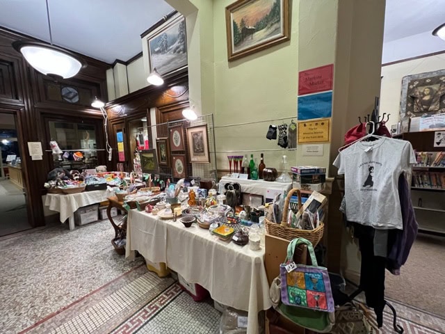 a room with tables displaying various items for sale, including dishes, bottles, baskets, books, and shirts; artwork and signs are on the walls.