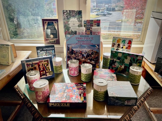 a selection of puzzles and candles arranged on a glass table in front of a window with a neighborhood view outside.