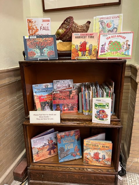 a wooden bookshelf displays fall themed children's books, including titles about autumn, apples, and harvest, with a ceramic turkey on top and a sign inviting people to check out the books.