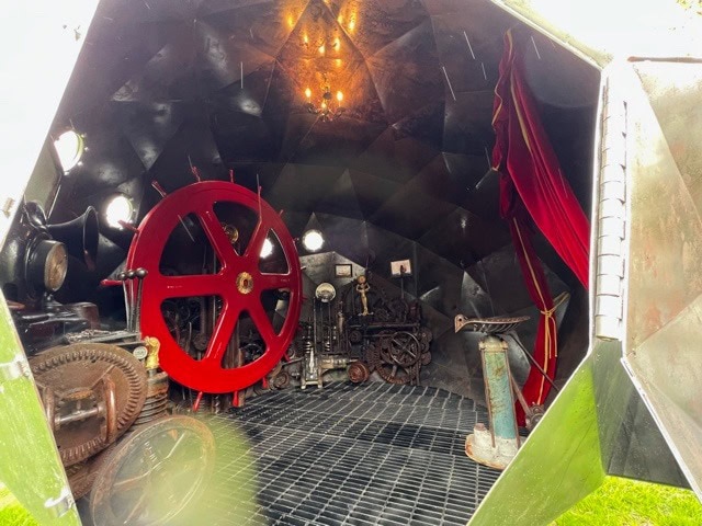 interior of a metallic, dome shaped room with large red machinery wheel, various mechanical gears, pipes, and tools; red curtain on the right side, with overhead light.