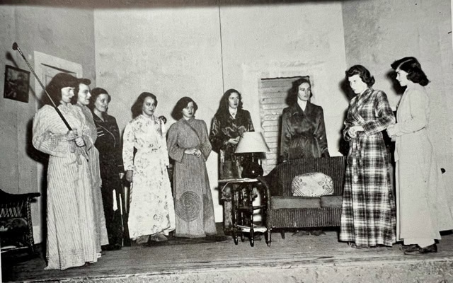 eight women stand on a stage in vintage attire, gathered around a small table and a lamp, appearing to participate in a dramatic performance or rehearsal.