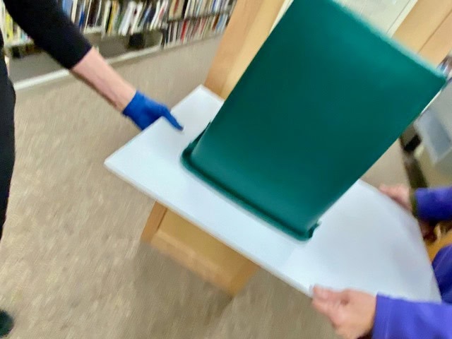 two people move a large green bin on a white board inside a room lined with bookshelves.