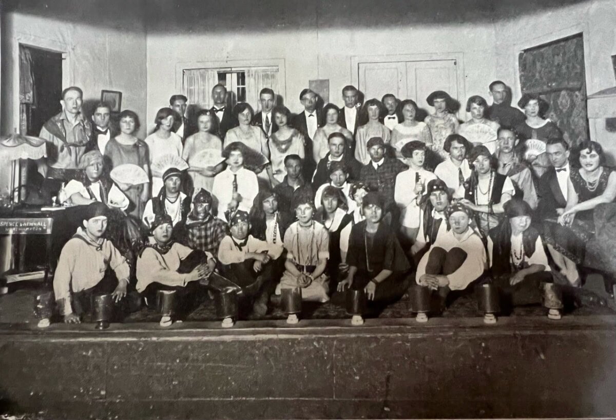 a large group of men and women pose indoors for a formal portrait, with several people in costume seated in the front row and others standing behind them.