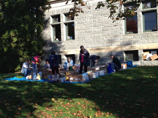 a group of people organize and sort boxes on a blue tarp outside a stone building on a sunny day.
