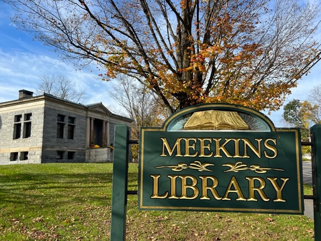 a green sign reading “meekins library” stands in front of a stone building with a tree shedding autumn leaves in the background.