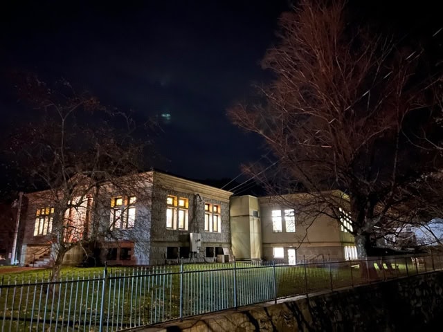 a large building with illuminated windows at night, surrounded by leafless trees and a metal fence, under a dark, cloudy sky.