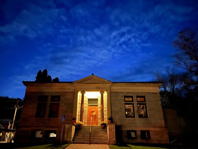 a stone building with columns is lit from the entrance at night, with a dark blue sky and scattered clouds in the background.