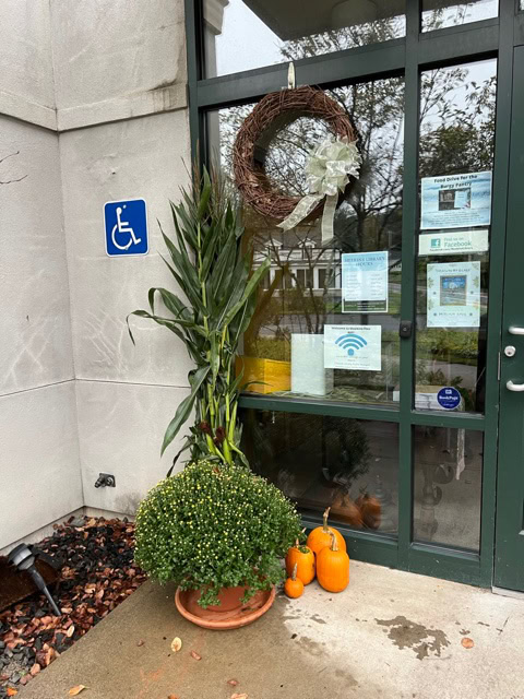 a glass door decorated with a large wreath and several notices, next to a potted plant and pumpkins, with a handicap accessible sign on the adjacent wall.