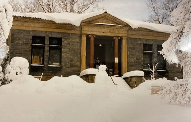 a small stone building with four columns at the entrance is surrounded by heavy snow, with snow covering the roof, steps, and trees in the foreground.