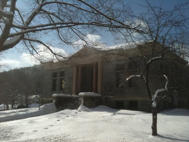 a stone building with columns at the entrance is surrounded by snow covered ground and bare trees under a clear, sunny sky.