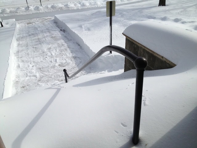 a freshly shoveled path leads away from a staircase buried under deep snow, with a handrail partially visible above the snowdrift.