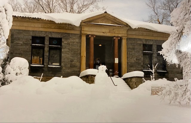 a stone building with four columns at the entrance is surrounded by deep snow, with a snowman in front and snow covered trees in the background.