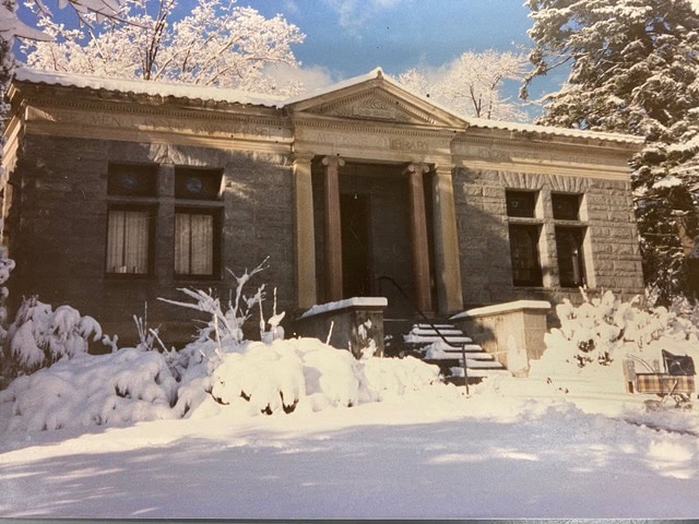 a small, stone library building with columns is covered in snow; trees and bushes in the surrounding yard are also blanketed in snow.