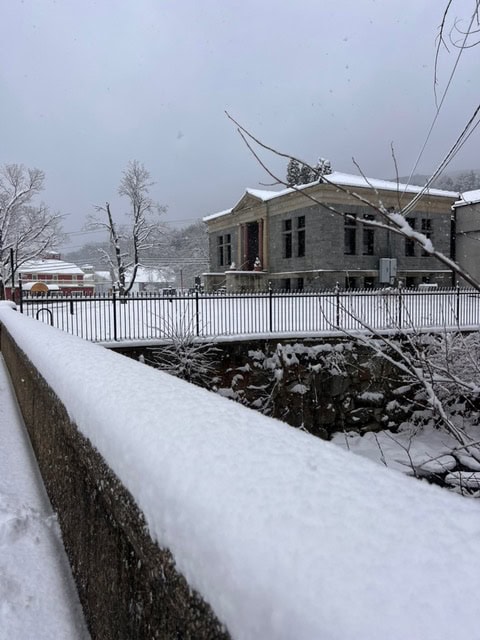 a stone building and a bridge are shown on a snowy day, with a layer of snow covering the ground, trees, and railing in the foreground.