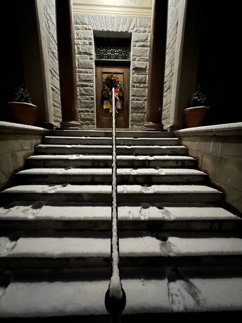 stone steps covered in snow lead up to a wooden door decorated with a holiday wreath, flanked by two columns and potted plants at night.