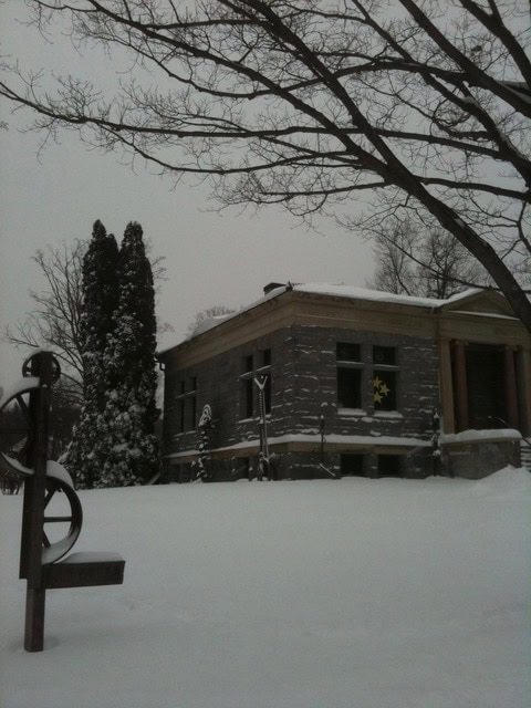 a gray stone building with columns stands in a snowy landscape, with bare tree branches overhead and a metal sculpture in the foreground.