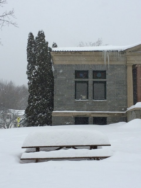 a snow covered picnic table sits in front of a stone building with icicles hanging from the roof on a snowy, overcast day.
