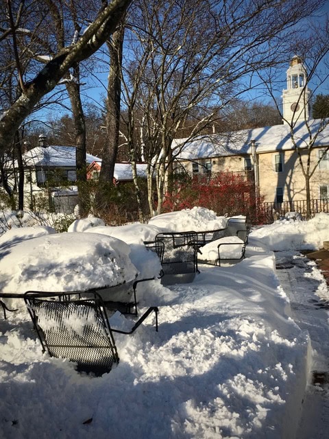 outdoor metal tables and chairs covered in deep snow on a patio, with bare trees and buildings in the background under a clear blue sky.