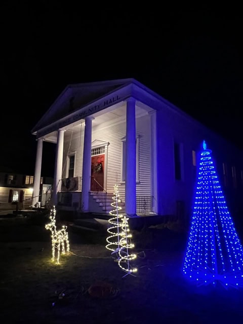 a white building with columns is decorated with holiday lights, including a lit reindeer, a yellow tree, and a blue cone shaped tree in the foreground at night.